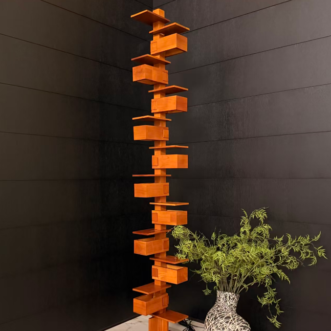 A tall, modern wooden shelving tower with multiple staggered square shelves in a warm orange-brown finish, standing on a marble floor against a dark paneled wall next to a patterned vase with green branches.