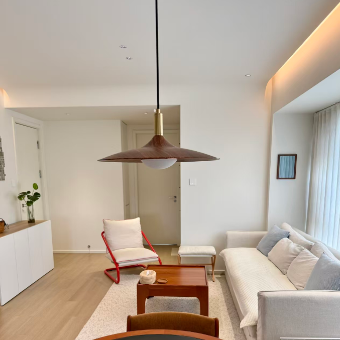 A modern living room interior featuring a light beige sofa with blue-gray pillows, a red metal accent chair, a wooden coffee table, and a large pendant light hanging above. The room has light wood floors, white walls, and a minimalist aesthetic.