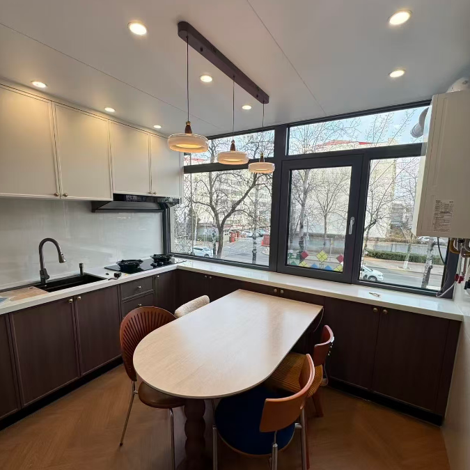 A modern kitchen and dining area with a light wood oval table surrounded by four chairs, set in a spacious kitchen with dark brown cabinets, white upper cabinets, and large windows.