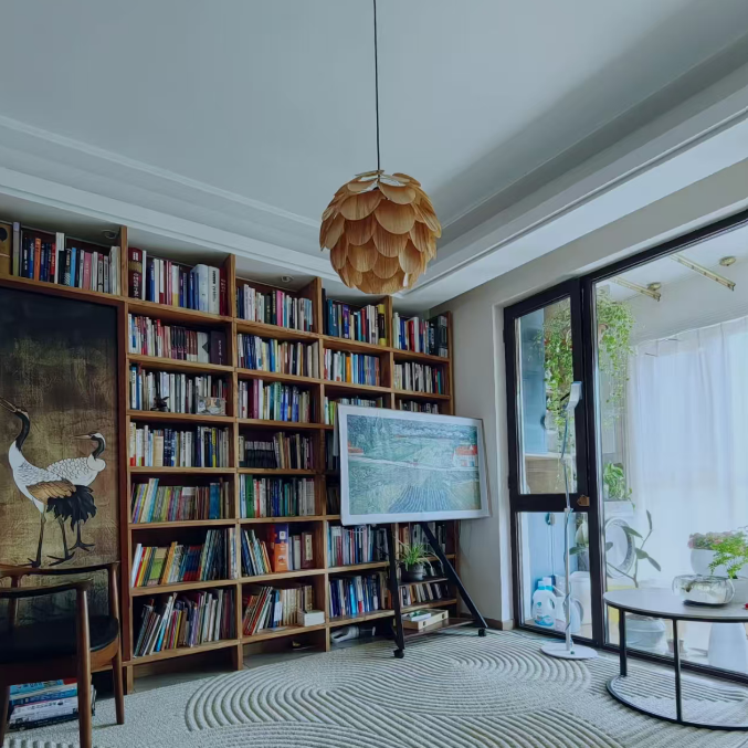 A modern living room featuring a large wooden bookshelf filled with books, a hanging paper lantern pendant light, a patterned gray rug, and a glass sliding door leading to a sunlit patio.