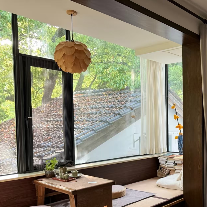 A cozy Japanese-style indoor seating nook with a low wooden table, round cushions, a hanging paper lantern, and large windows overlooking a tiled roof and trees.