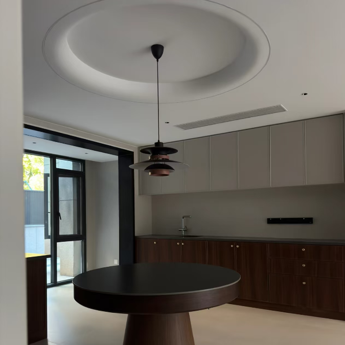 A modern round dining table with a dark top and brown tapered pedestal base, set in a minimalist dining room with a matching dark pendant light and kitchen cabinets in the background.