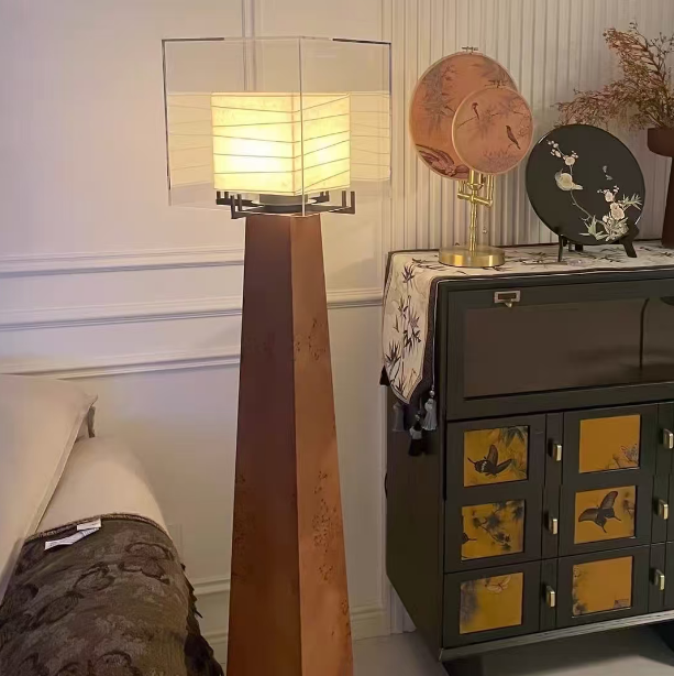 A tall floor lamp with a rectangular clear shade and a tapered wooden base, glowing with warm light in a styled interior next to a dark cabinet.