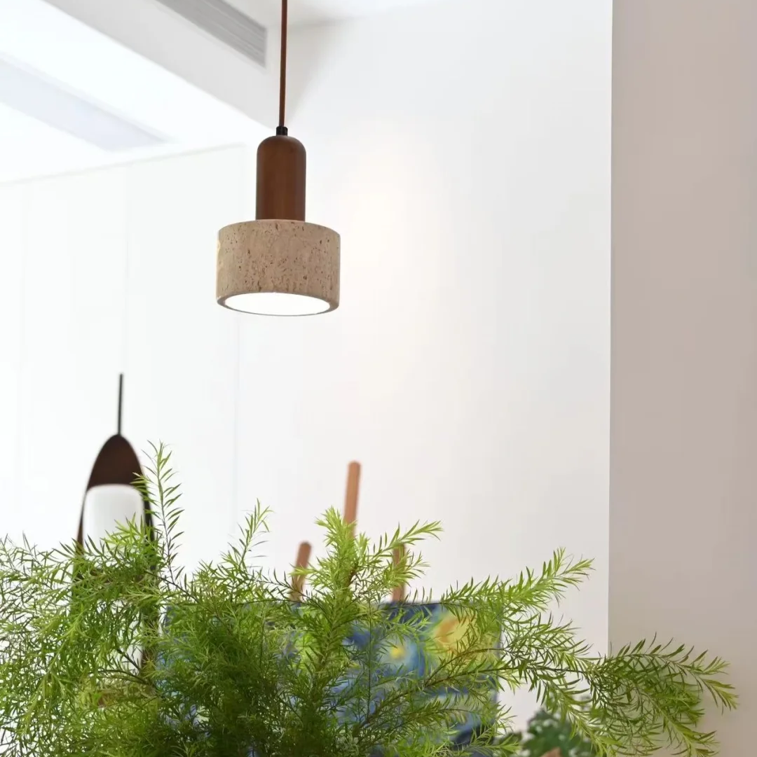 A modern cylindrical pendant light with a beige textured fabric shade and a dark brown wooden top, hanging in a bright, minimalist room above a wooden table with a green potted plant.