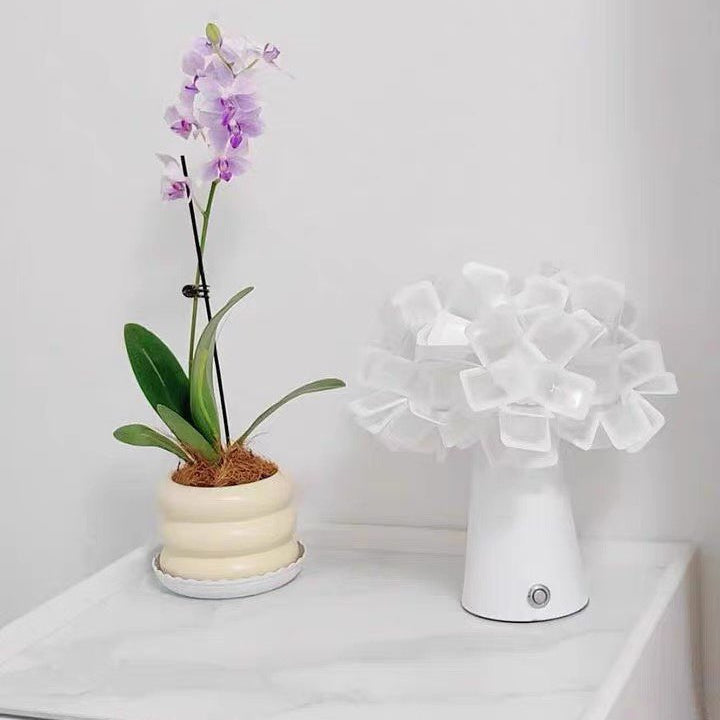 Geometric blossom table lamp with white base and white glowing cube petal shade.