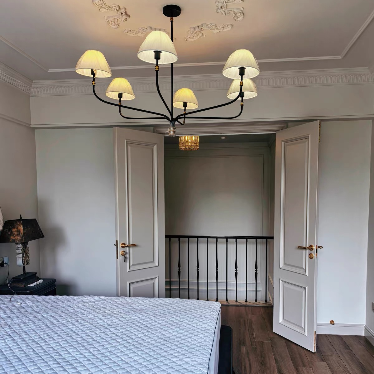 Cream fabric shade chandelier with black arms and gold finial centered above a bedroom bed.