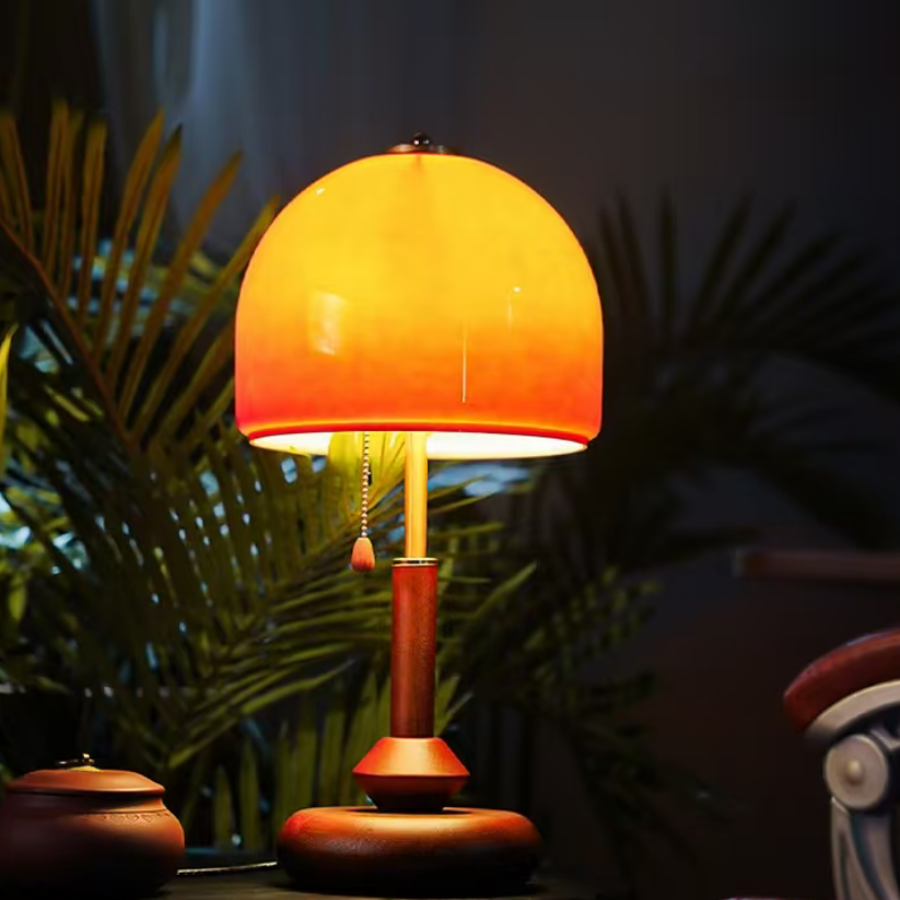A table lamp with a warm orange gradient glass dome shade and a reddish-brown wooden base, illuminated in a cozy setting with dark green foliage in the background.