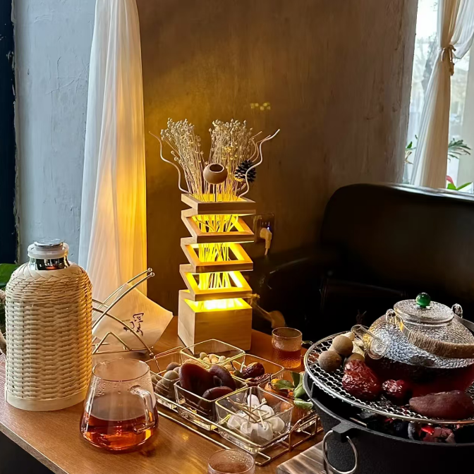 A warm, cozy interior scene with a glowing wooden tiered lamp featuring dried grasses and a small bird, set on a table alongside tea glasses, a woven bottle, and a teapot over a stove in a dimly lit room.