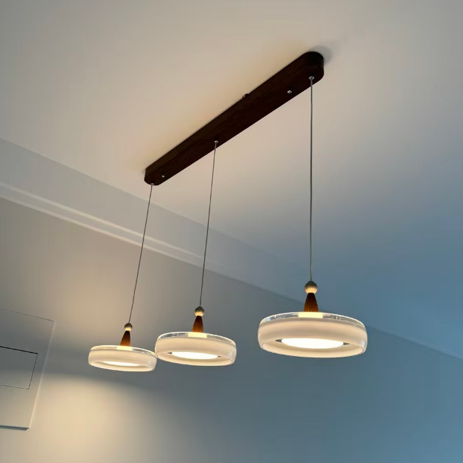 A modern three-light linear pendant chandelier with frosted glass circular shades and warm wood accents, suspended from a dark wood ceiling bar in a room with light gray walls.