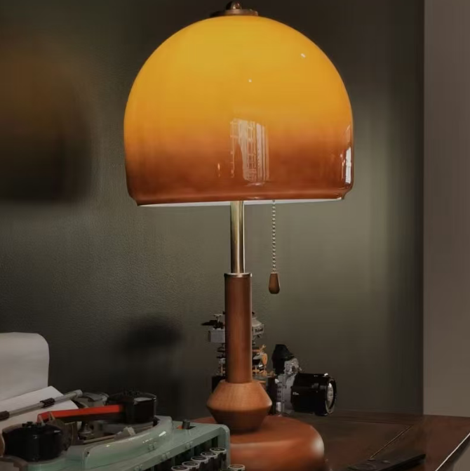 A vintage-style table lamp with a warm orange-to-brown gradient glass dome shade, a brown wooden base, and a metal stem, shown lit on a dark wooden desk next to a typewriter.