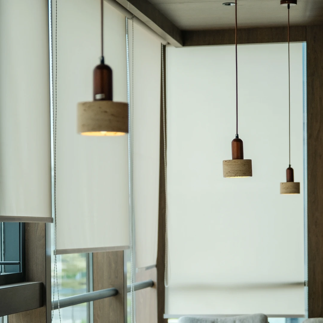 A modern dining or lounge interior with light gray upholstered chairs, wooden tables, large windows with white roller blinds, and three hanging pendant lights with cylindrical shades.