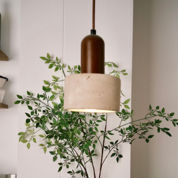 A modern pendant light with a textured beige concrete drum shade and a dark brown cylindrical top, hanging in a minimalist room next to a green leafy plant in a ceramic vase.