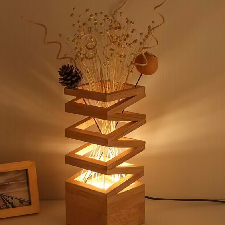 A wooden spiral staircase table lamp with built-in warm white LED lights, styled with dried botanicals and a pinecone on a beige background.