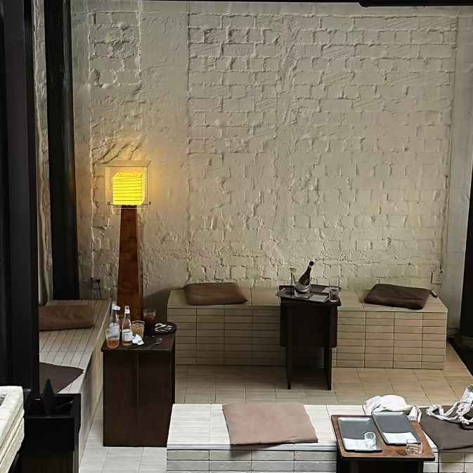 A spa-like interior with a textured cream brick wall, built-in seating with folded towels, a glowing floor lamp, and small wooden tables holding bottles, glasses, and trays.