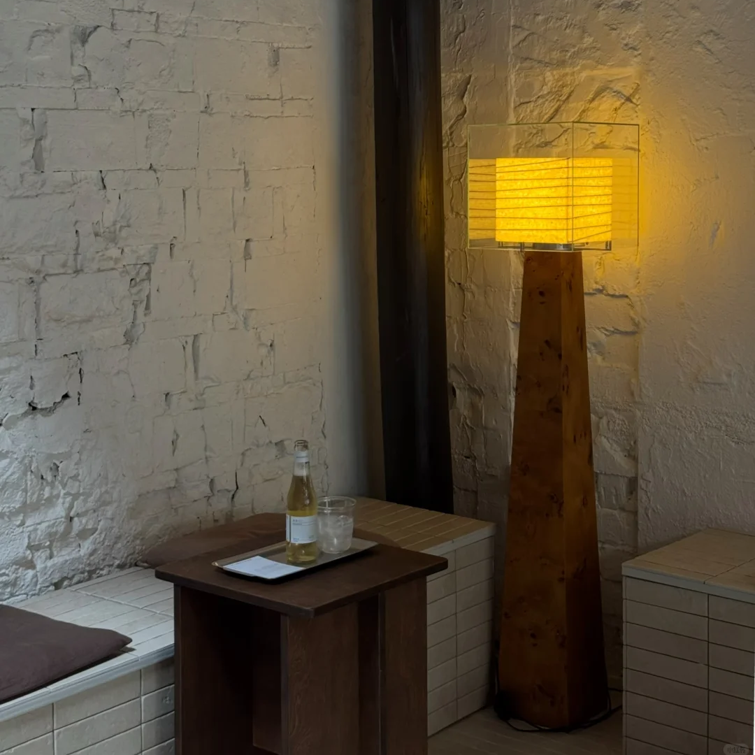 A tall floor lamp with a glowing warm yellow rectangular shade and a dark wood base, standing in a corner of a room with textured white brick walls. A small wooden side table with a bottle and glass sits nearby.