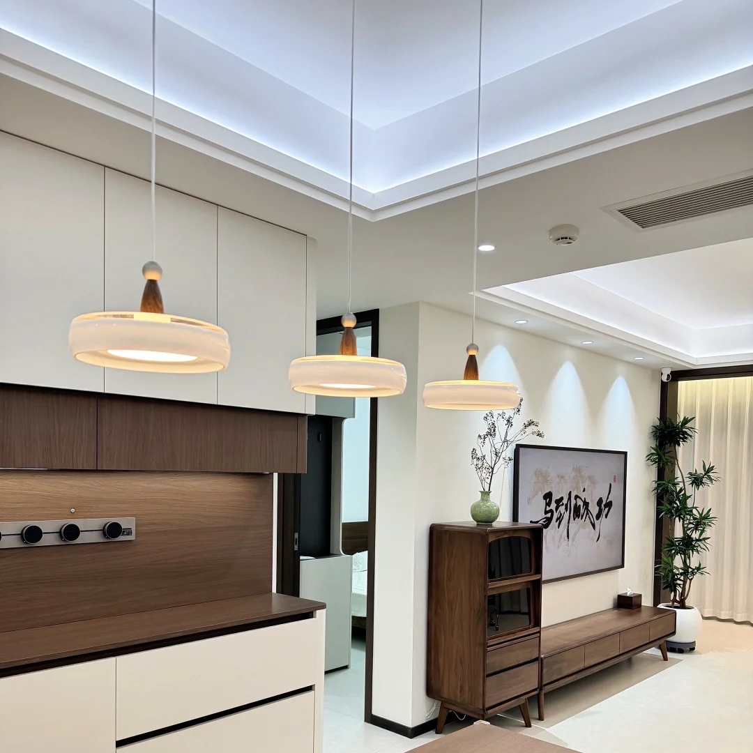 Modern dining area featuring a rectangular wooden table in the foreground, three matching pendant lights hanging from the ceiling, and a contemporary kitchen cabinetry unit in the background.