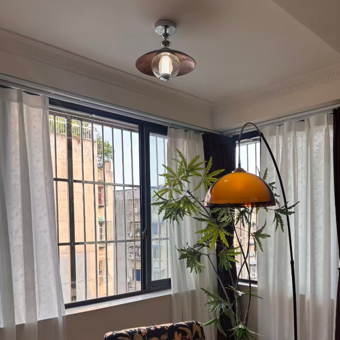 A modern ceiling fan with a bronze metal cupola and a single exposed bulb, shown installed in a real room with white curtains, a window view, a floor lamp, and a chair.
