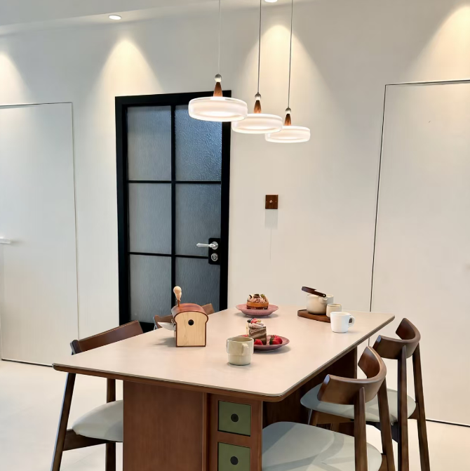 A mid-century modern dining table set in a bright, minimalist dining room. The table features a light beige top, a brown wooden pedestal base with four green drawer fronts, and is surrounded by four matching wooden chairs with light gray upholstered seats.
