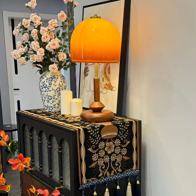 A decorative black and gold embroidered table runner with tassels draped over a cabinet, styled with a glowing orange table lamp, white candles, and a blue-and-white floral vase in a home interior.