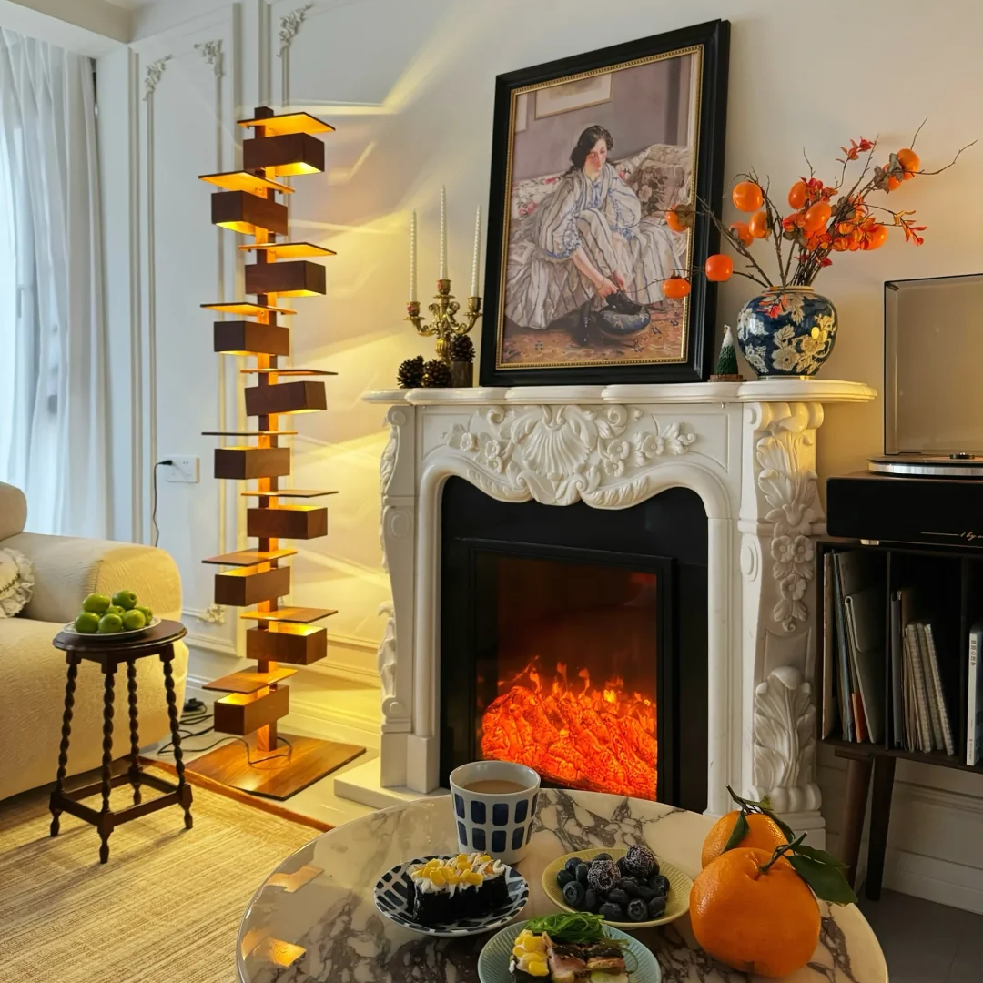 A styled interior scene featuring a white carved fireplace with an electric fire burning inside, a framed portrait on the mantel, a tall sculptural floor lamp, and a marble coffee table in the foreground.