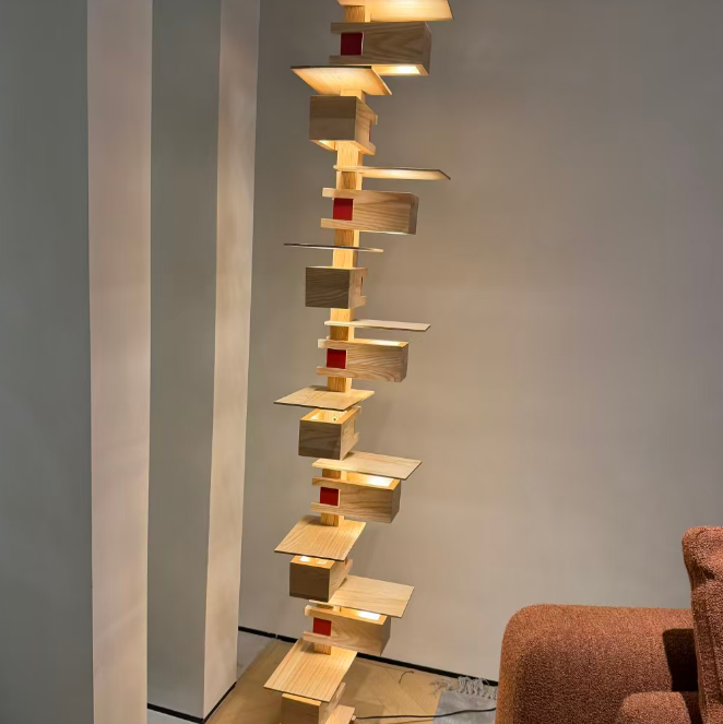A modern abstract wooden floor lamp with stacked rectangular wood blocks and integrated warm lighting, shown in a minimalist room setting next to a brown armchair.