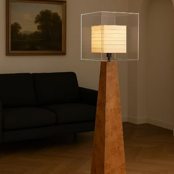 A modern floor lamp with a tapered wood-grain base and a clear square acrylic shade, lit with a warm white horizontal pleated shade, standing in a living room next to a black sofa.
