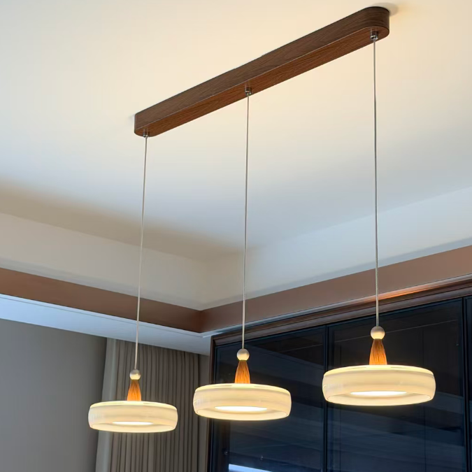 Three-light linear ceiling pendant chandelier with a dark wood bar, thin metal rods, and round white shades with amber-colored accents, shown installed in a modern room with a window and curtains.