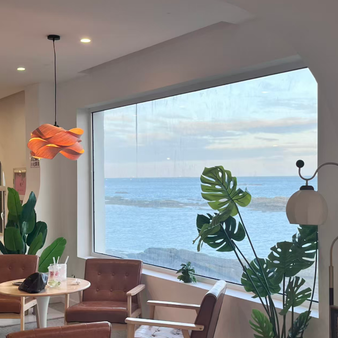 A modern living room corner with a large window overlooking the ocean, furnished with brown leather armchairs, a small round table, a hanging orange flower-shaped pendant light, and tall green monstera plants.