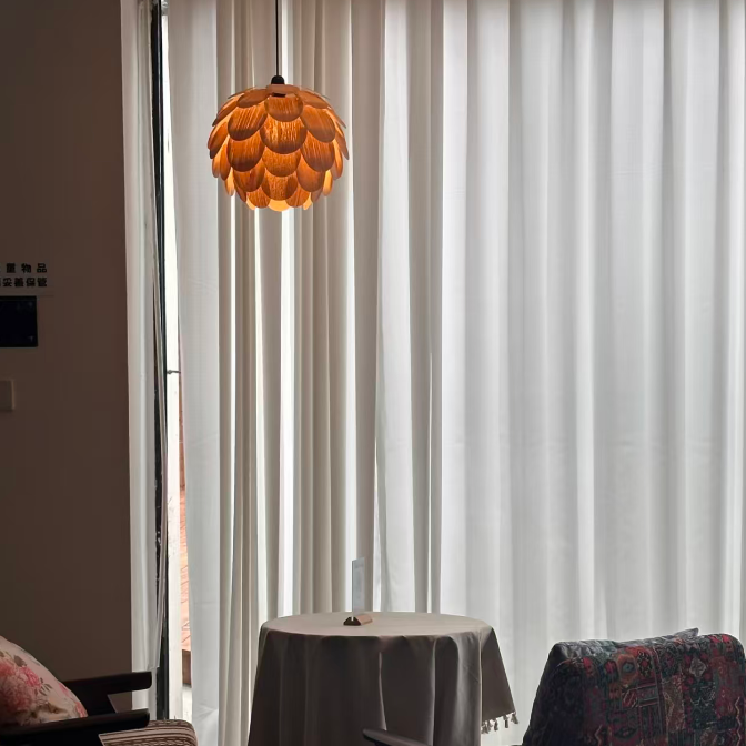 A modern pendant light with a textured, layered orange-gold flower or lotus-shaped shade hangs in a styled room with white sheer curtains, a small round table, and two chairs.