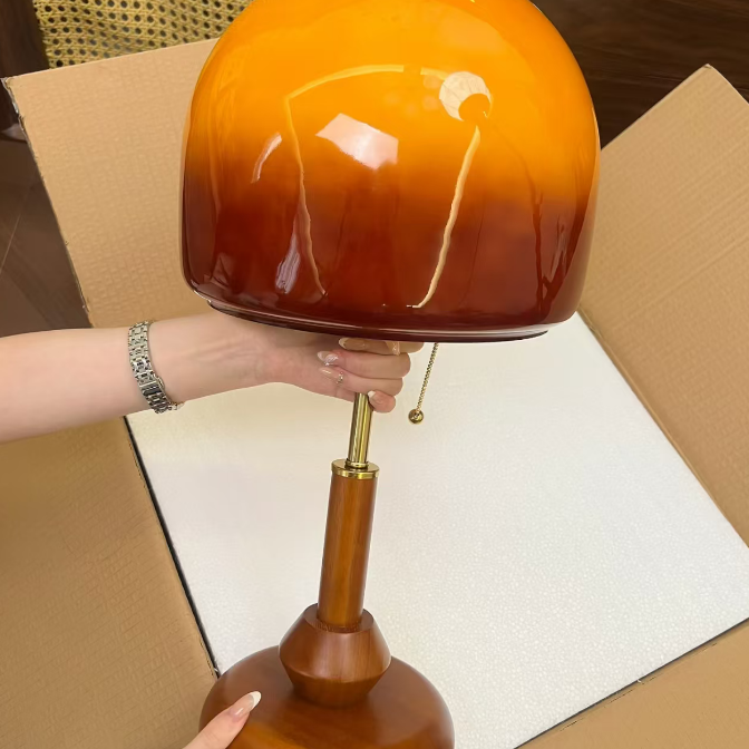A glossy table lamp with a spherical shade featuring a warm orange-to-brown gradient, supported by a polished wooden base and brass-toned stem, being held by a person inside an open cardboard box.
