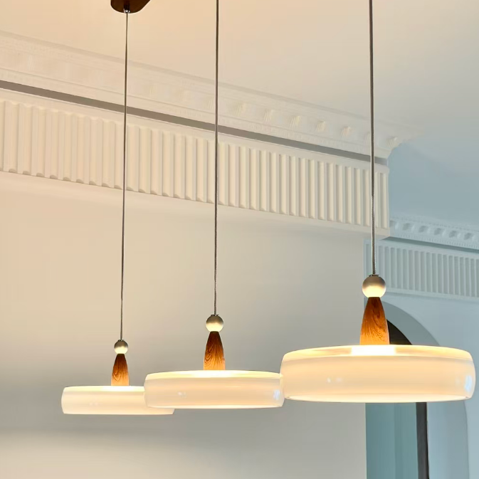 Three modern pendant lights with circular white shades and warm wood-toned accents hanging from thin black cords in an indoor setting.