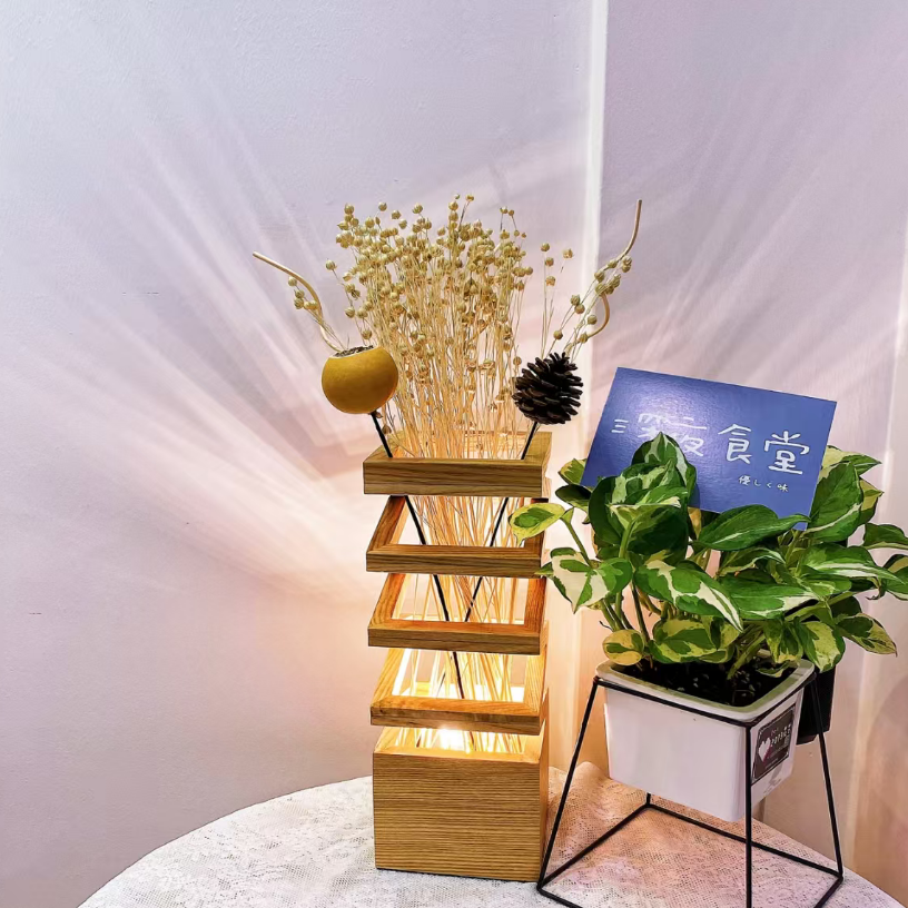 A wooden tiered candle holder with warm white LED lights inside, styled with dried flowers and a small yellow ornament, next to a potted green plant on a white table against a draped fabric background.