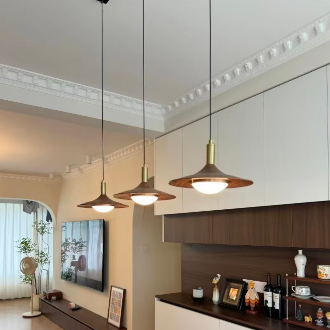 Three modern pendant lights with conical bronze shades and warm white glowing bulbs, hanging in a dining or kitchen area with white and dark wood cabinetry.