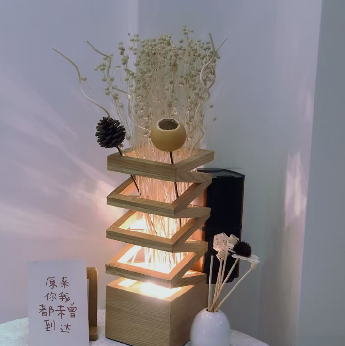 A modern wooden tiered table lamp with warm LED lighting, styled with dried flowers, a pinecone, and a small card on a tabletop against a neutral wall.