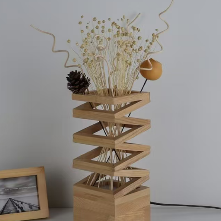 A modern wooden geometric table lamp with a zigzag staircase design, filled with dried grasses, small gold beads, a pinecone, and a small orange decorative piece. The lamp is set on a light gray surface against a neutral gray wall, with a small picture frame partially visible on the left.