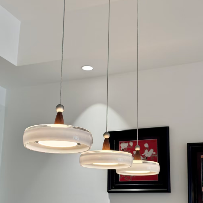Three modern pendant lights with frosted white circular shades and small brown wooden accents, hanging from thin cords in a bright room with white walls and framed wall art.