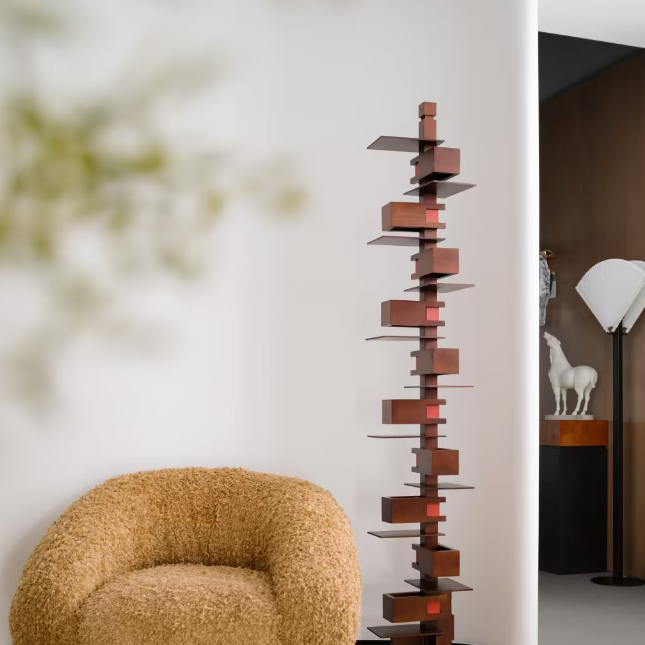 A tall, modern wooden geometric floor lamp with multiple stacked rectangular shelves stands in a minimalist room next to a textured mustard yellow armchair.
