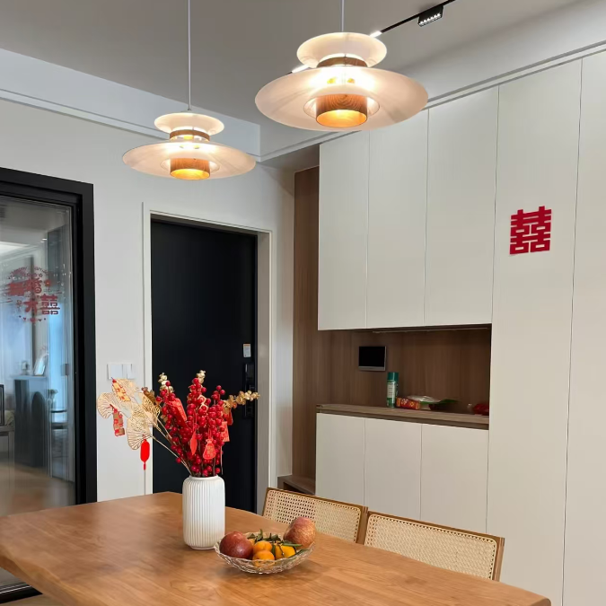 A modern dining room featuring a large, rustic rectangular wooden table with a live-edge style, paired with woven rattan chairs. The table is decorated with a white vase of red artificial flowers and a bowl of fruit.