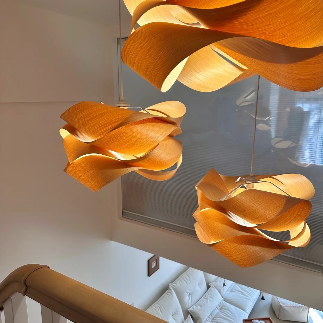 Three modern pendant lights with sculptural, flowing wooden leaf-like shapes hanging in a bright, contemporary home interior above a staircase and living area.