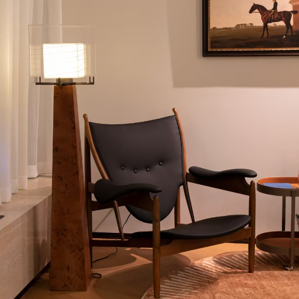 A mid-century modern lounge chair with black tufted upholstery and a curved wooden frame, styled in a warm-toned room next to a wooden floor lamp and a framed equestrian painting.