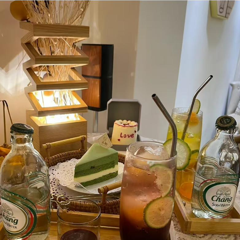 A cozy cafe table setting featuring a glass of iced dark cola with lime slices, a green slice cake, and two bottles of Chang soft drink on a wooden tray, with a decorative wooden light fixture in the background.