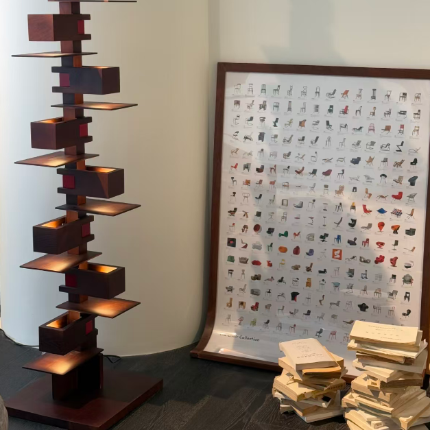 A tall, modern wooden floor lamp with a staggered, geometric cube design and integrated warm lighting stands next to a large framed reference chart of lamp shapes on a white wall. A stack of books sits on the floor nearby.