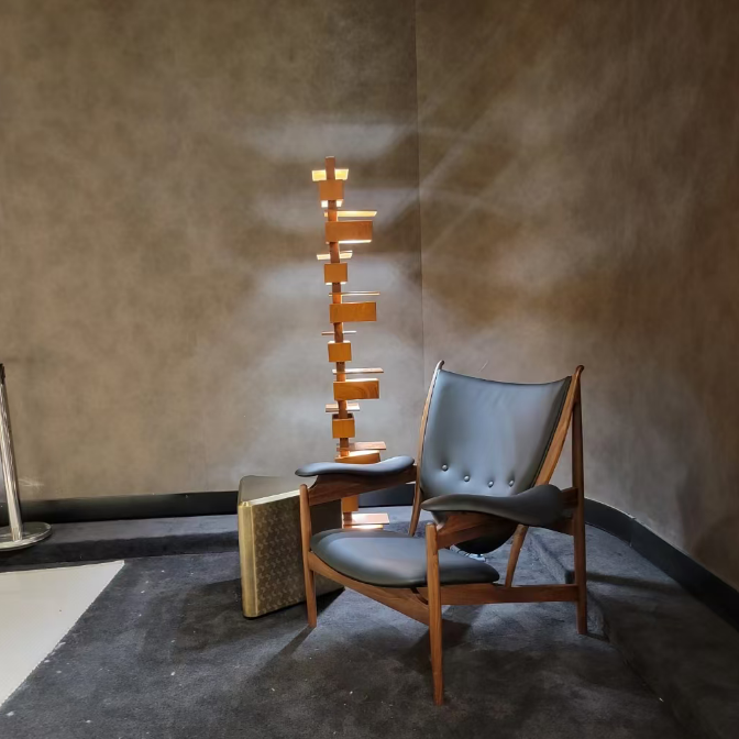 A mid-century modern wooden armchair with dark gray leather upholstery, positioned next to a tall, minimalist wooden block light fixture and a small patterned side table against a textured brown plaster wall.