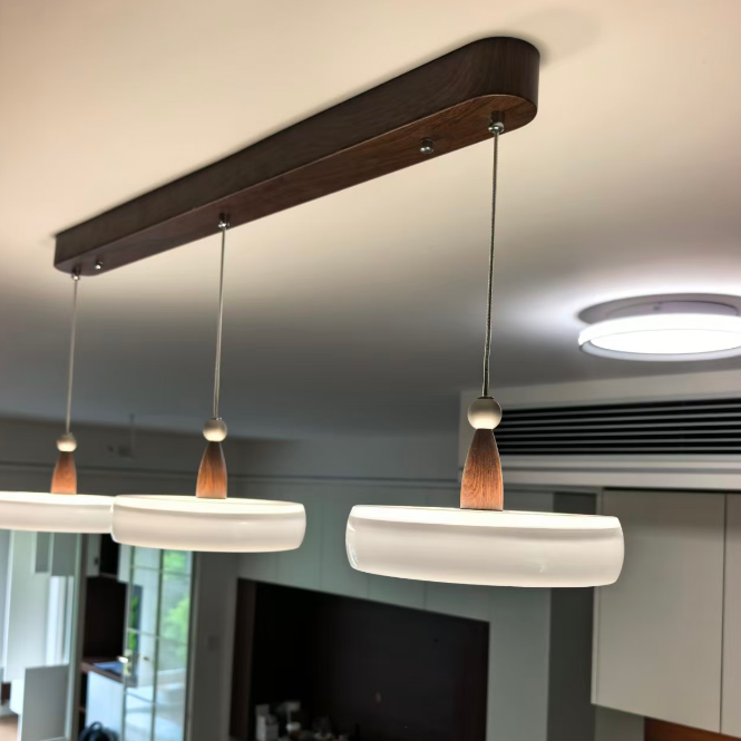 A modern linear pendant light fixture with a dark wood ceiling mount, three white cylindrical shades, and small wooden finials, shown installed in a real interior room.